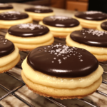 A close-up of Boston cream pie cookies, featuring soft cookies filled with creamy custard and topped with glossy chocolate ganache, arranged on a plate.